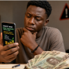A Ghanaian looking at a smartphone displaying a suspicious investment advert while holding Ghana cedi notes, illustrating the dangers of get rich quick scams