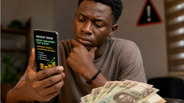 A Ghanaian looking at a smartphone displaying a suspicious investment advert while holding Ghana cedi notes, illustrating the dangers of get rich quick scams